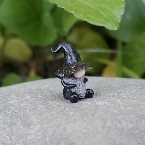 May include: A small black gnome figurine with a black pointed hat and a gray beard. The gnome is sitting on a gray rock.