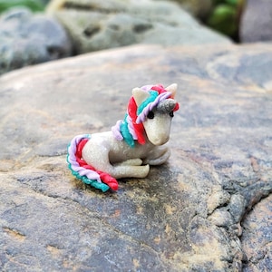 May include: A small, white unicorn figurine with a rainbow mane and tail. The unicorn is sitting on a gray rock.