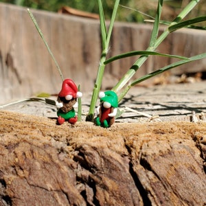 May include: Two small gnome figurines made of clay. One gnome is wearing a red hat and the other is wearing a green hat. Both gnomes are holding a small brown object in their hands. The gnomes are sitting on a brown wooden surface.