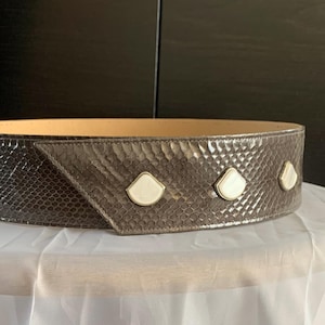 Może przedstawiać: Szeroki, brązowy pasek ze wzorem skóry węża. Pasek posiada trzy białe, ozdoby w kształcie muszli. Pasek jest prezentowany na białej powierzchni.