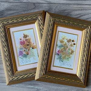 Pode incluir: Duas peças de arte de flores prensadas emolduradas com molduras douradas. As molduras têm um design de corda decorativo. As peças de arte estão sobre um fundo de madeira marrom claro.