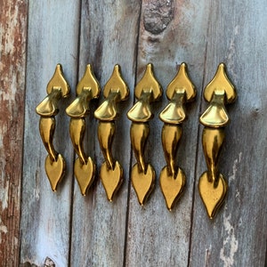 以下が含まれることがあります： 装飾的なデザインが施された、金色のハート型のキャビネットプルが7つ。プルは木製の背景に一列に並んでいます。