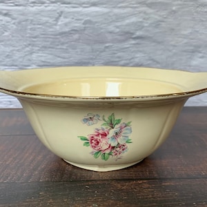 Puede incluir: Un tazón de cerámica de color crema con un diseño floral. El tazón tiene un borde festoneado y un borde dorado. El diseño floral presenta flores rosas, azules y verdes.