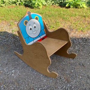 以下が含まれることがあります： 背もたれにトーマス機関車のキャラクターが描かれた、子供用の木製ロッキングチェア。椅子は茶色で、青、赤、黄色のトーマス機関車のデザインが施されています。