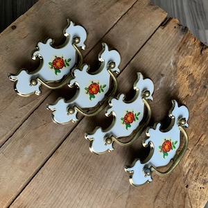 May include: Four white ceramic drawer pulls with gold accents and red floral designs. The pulls have a decorative, ornate shape.