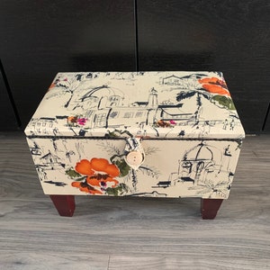 Peut inclure: Une boîte de rangement blanche avec un imprimé floral et architectural. La boîte a une fermeture à bouton et repose sur quatre pieds en bois.