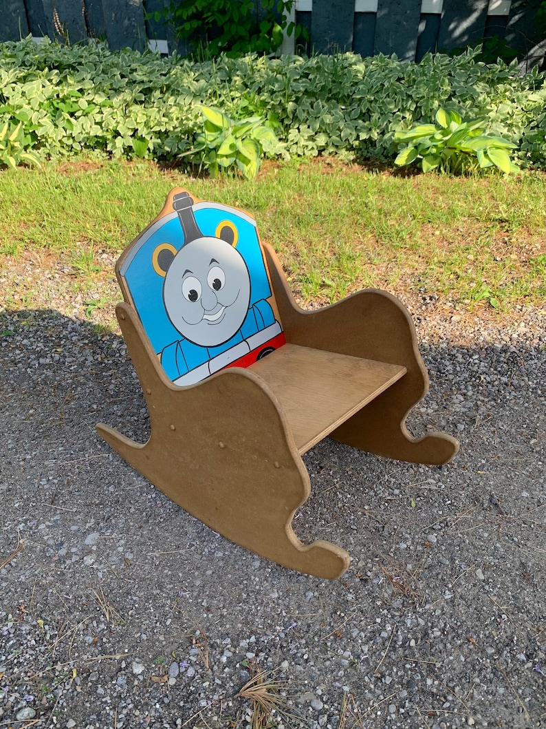 以下が含まれることがあります： 背もたれにトーマス機関車のキャラクターが描かれた、子供用の木製ロッキングチェア。椅子は茶色で、青、赤、黄色のトーマス機関車のデザインが施されています。