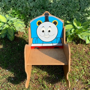以下が含まれることがあります： トーマス機関車の形をした木製ロッキングチェア。椅子は青、赤、黒に塗られており、背もたれにはトーマスの漫画の顔が描かれています。椅子は緑の芝生の上に置かれています。