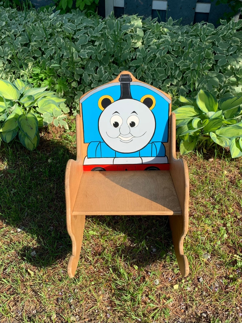 以下が含まれることがあります： トーマス機関車の形をした木製ロッキングチェア。椅子は青、赤、黒に塗られており、背もたれにはトーマスの漫画の顔が描かれています。椅子は緑の芝生の上に置かれています。