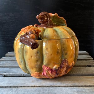 May include: A ceramic pumpkin-shaped serving bowl with a lid. The bowl is decorated with fall leaves and acorns in shades of brown, orange, and green.