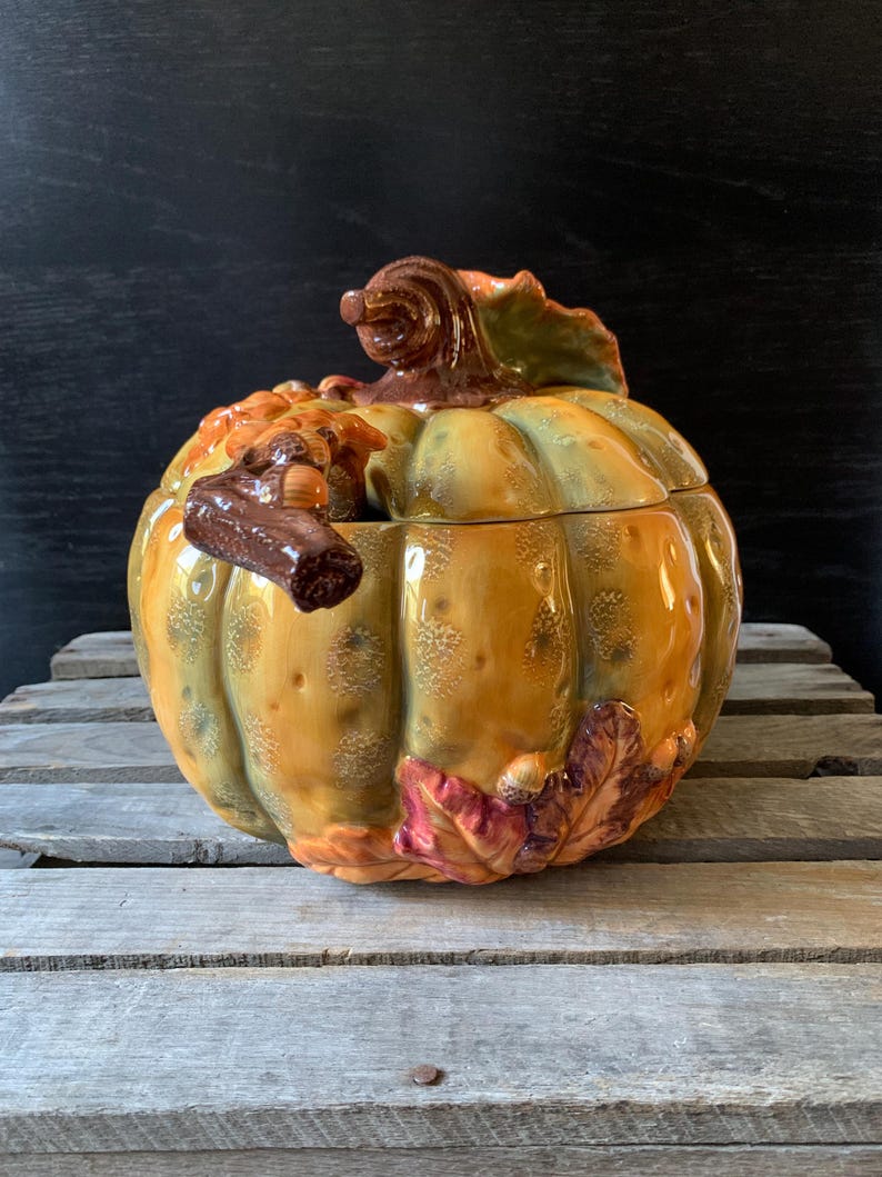 May include: A ceramic pumpkin-shaped serving bowl with a lid. The bowl is decorated with fall leaves and acorns in shades of brown, orange, and green.