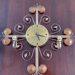 Op de afbeelding: Een goudkleurige wandklok met een zwarte wijzerplaat en zwarte wijzers. De klok is gemonteerd op een decoratief metalen frame met goudkleurige bladeren en kandelaars. Het frame is bevestigd aan een houten muur.