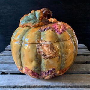 May include: A ceramic pumpkin-shaped container with a lid, decorated with fall leaves in shades of green, brown, red, and purple. The container is a light yellow color and has a textured surface.