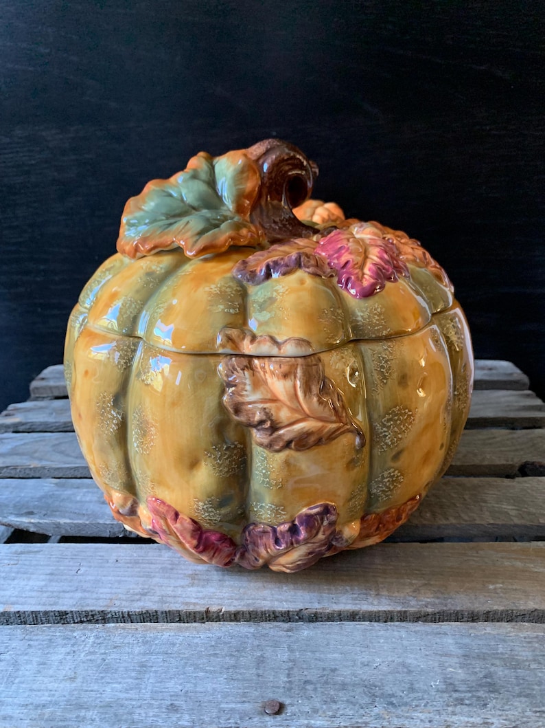 May include: A ceramic pumpkin-shaped container with a lid, decorated with fall leaves in shades of green, brown, red, and purple. The container is a light yellow color and has a textured surface.