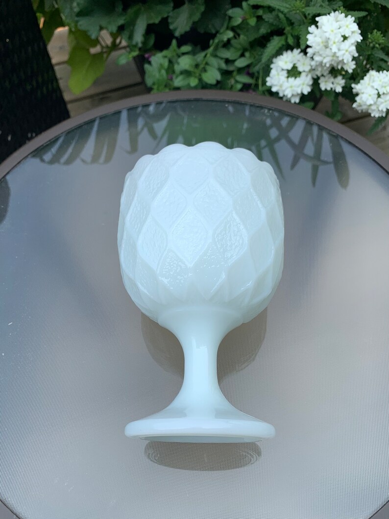 K&ouml;nnte beinhalten: Eine wei&szlig;e Milchglasvase mit einem strukturierten, rautenf&ouml;rmigen Muster. Die Vase hat einen breiten, runden Boden und einen hohen, schlanken Stiel.