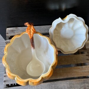 May include: A ceramic pumpkin-shaped serving bowl with a lid and a ladle. The bowl is decorated with a brown stem and a leaf, and the lid has a scalloped edge.