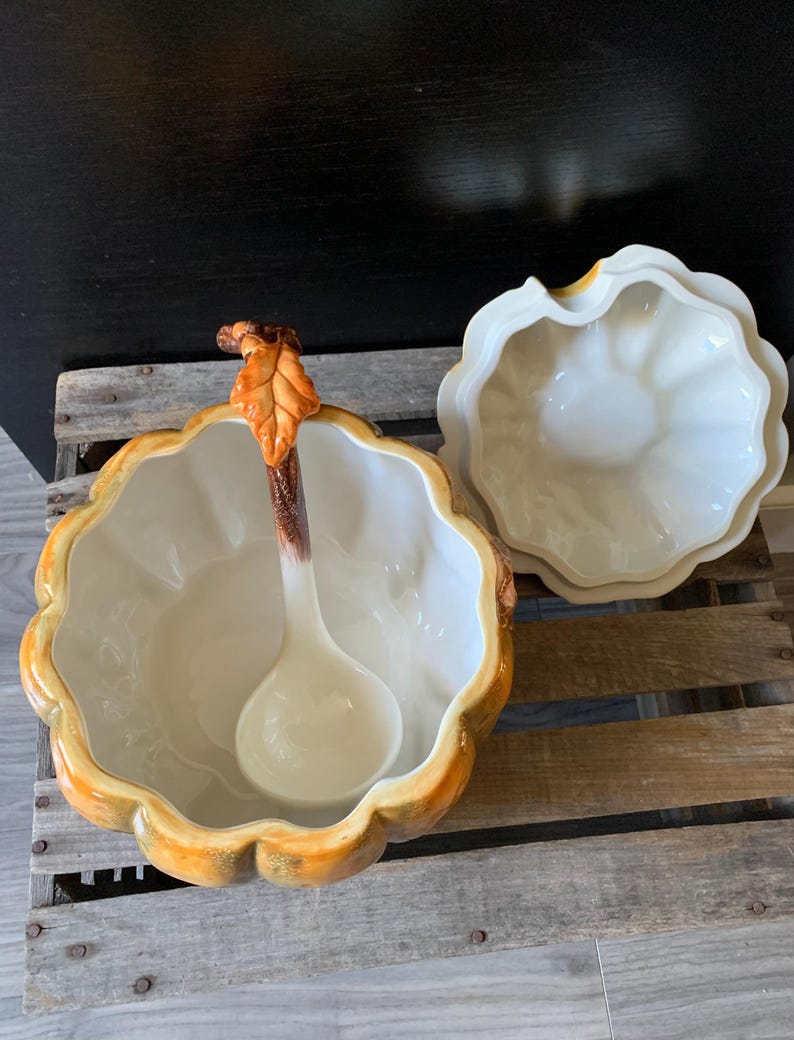 May include: A ceramic pumpkin-shaped serving bowl with a lid and a ladle. The bowl is decorated with a brown stem and a leaf, and the lid has a scalloped edge.