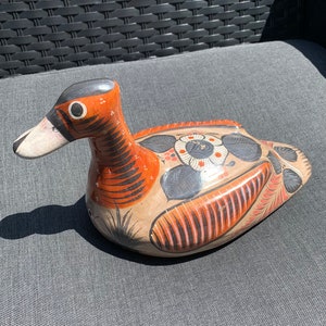 Può includere: Figurina di anatra in ceramica dipinta a mano con dettagli arancioni, neri e bianchi. L'anatra è sdraiata con la testa girata di lato.