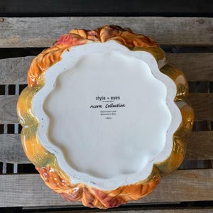 May include: A ceramic pumpkin-shaped serving dish with a fall leaf design. The dish is white with brown, orange, and green accents. The bottom of the dish has the text "style - eyes by home edit Acorn Collection Dishwasher and Microwave Safe China"