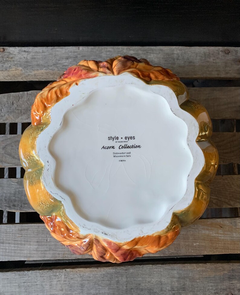 May include: A ceramic pumpkin-shaped serving dish with a fall leaf design. The dish is white with brown, orange, and green accents. The bottom of the dish has the text "style - eyes by home edit Acorn Collection Dishwasher and Microwave Safe China"
