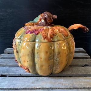 May include: A ceramic pumpkin-shaped serving bowl with a lid and a ladle. The bowl is decorated with fall leaves in shades of orange, red, and green. The bowl is sitting on a wooden surface.
