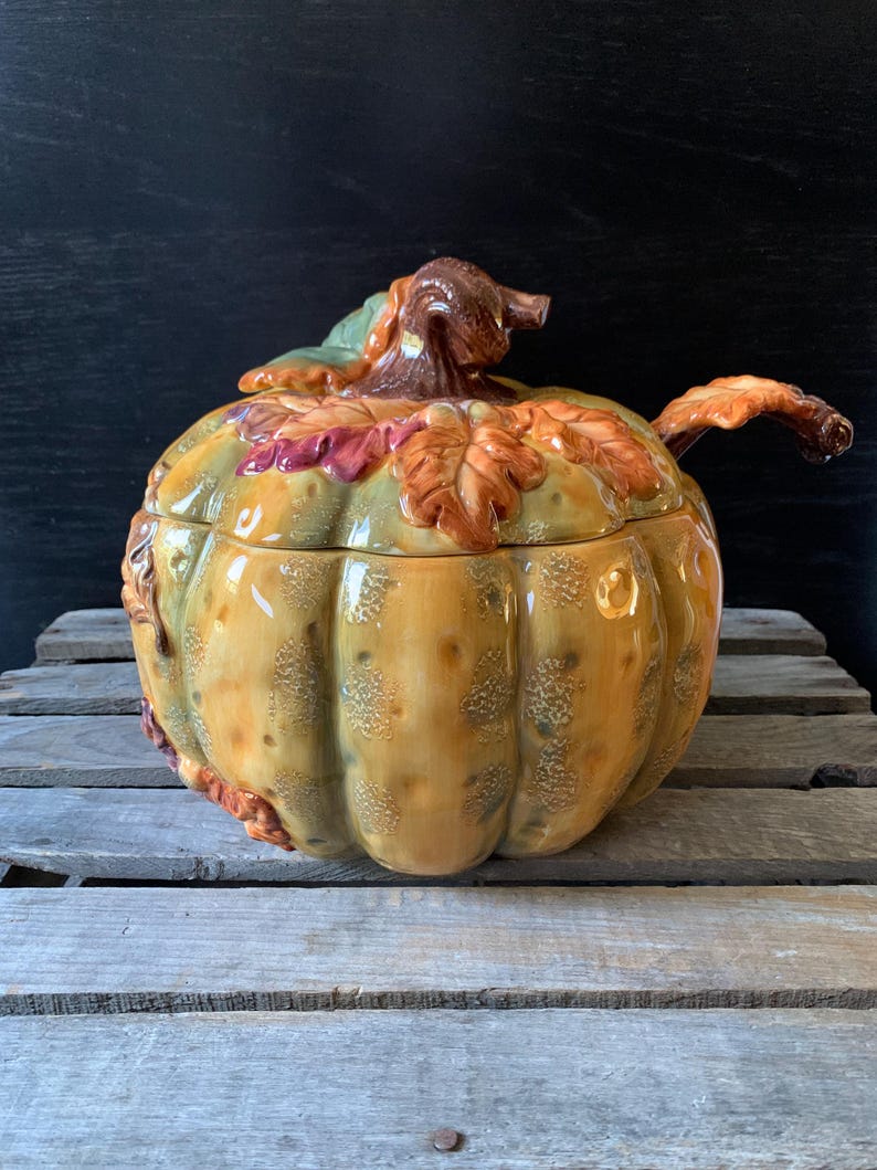 May include: A ceramic pumpkin-shaped serving bowl with a lid and a ladle. The bowl is decorated with fall leaves in shades of orange, red, and green. The bowl is sitting on a wooden surface.