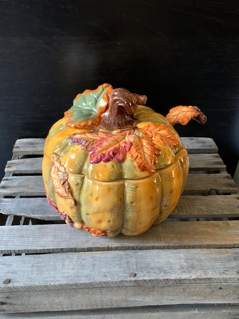 May include: A ceramic pumpkin-shaped serving bowl with a lid. The bowl is decorated with fall leaves in shades of red, orange, yellow, and green.