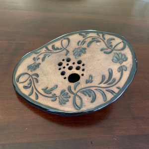 Peut inclure: Un plat en céramique brun avec un bord noir et un motif floral. Le plat a un trou au centre avec un anneau de petits trous autour.