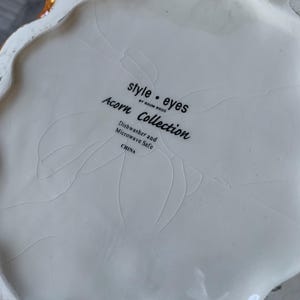 May include: White ceramic bottom of a decorative item with the text "style.eyes by Baum Bros. Acorn Collection Dishwasher and Microwave Safe CHINA"