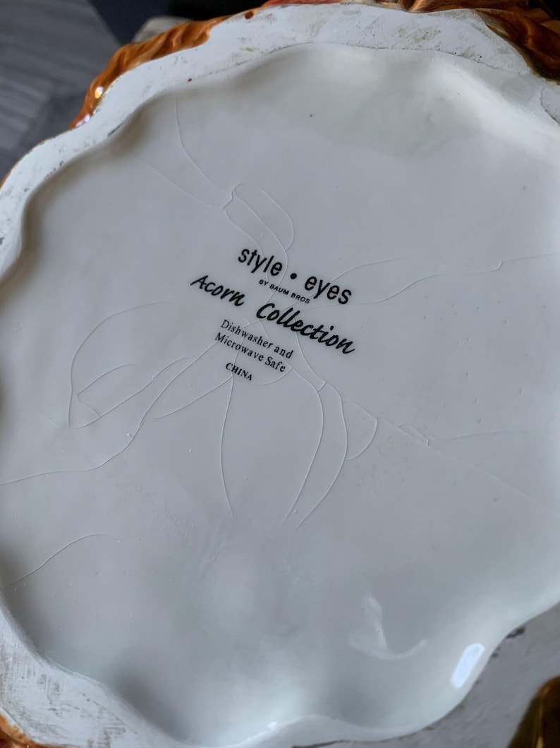 May include: White ceramic bottom of a decorative item with the text "style.eyes by Baum Bros. Acorn Collection Dishwasher and Microwave Safe CHINA"