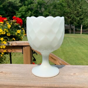K&ouml;nnte beinhalten: Eine wei&szlig;e Milchglas-Fu&szlig;schale mit einem gewellten Rand und einem Diamantenmuster. Die Schale steht auf einer Holzoberfl&auml;che.