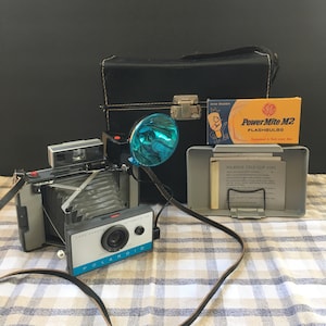 Puede incluir: Una cámara Polaroid Land Camera Automatic 104 vintage con cuerpo azul y plateado, un flash y una correa de cuero negra. La cámara está sobre un mantel a cuadros con una caja de flashes y una hoja de instrucciones de Polaroid Cold-Clip.