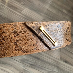 May include: A brown snakeskin belt with a gold metal buckle. The buckle is a simple, rectangular design with a straight line across the centre.