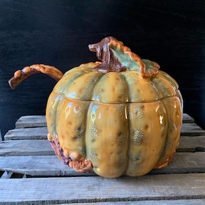 May include: A ceramic pumpkin-shaped serving bowl with a lid and a handle. The bowl is decorated with brown, green, and yellow glaze, and has a realistic pumpkin design.