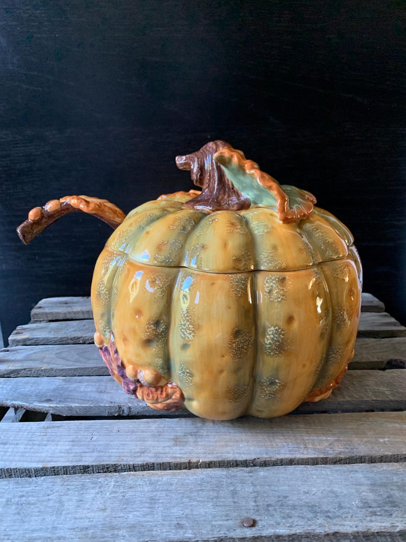 May include: A ceramic pumpkin-shaped serving bowl with a lid and a handle. The bowl is decorated with brown, green, and yellow glaze, and has a realistic pumpkin design.