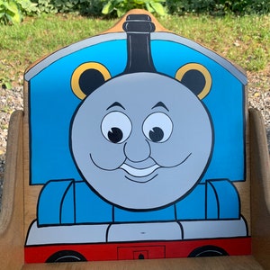 以下が含まれることがあります： 青いグレーのトーマス機関車のデザインが描かれた木製のおもちゃの椅子。椅子は赤いベースと黒い煙突があります。