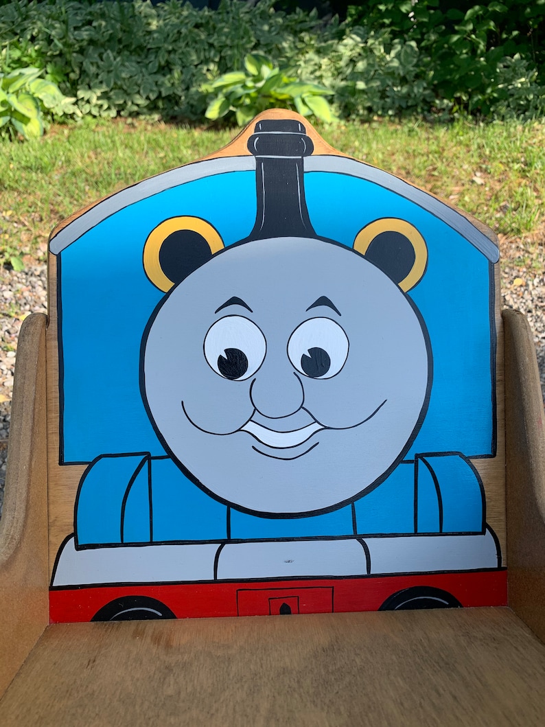 以下が含まれることがあります： 青いグレーのトーマス機関車のデザインが描かれた木製のおもちゃの椅子。椅子は赤いベースと黒い煙突があります。