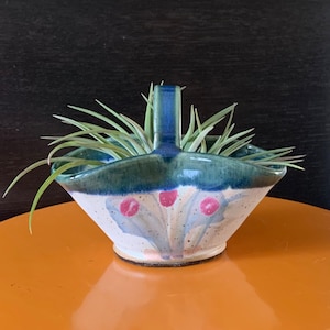 Puede incluir: Un pequeño macetero de cerámica blanca con un asa azul y diseños florales verdes y rosas. El macetero está lleno de una planta de aire verde.