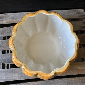 May include: A ceramic pumpkin-shaped bowl with a white interior and a yellow and gold exterior. The bowl is sitting on a wooden surface.