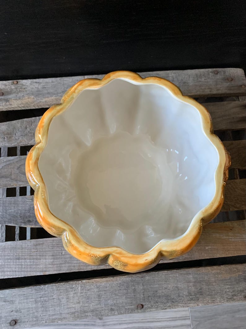 May include: A ceramic pumpkin-shaped bowl with a white interior and a yellow and gold exterior. The bowl is sitting on a wooden surface.
