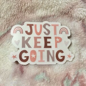 May include: A white sticker with the text "JUST KEEP GOING" in pink, brown, and gray letters. The sticker has a rainbow, hearts, and stars around the text.
