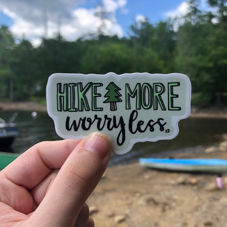 May include: A green and white sticker with the text "HIKE MORE worry less" and a small green tree icon. The sticker is being held by a hand in front of a blurry background of a lake and trees.