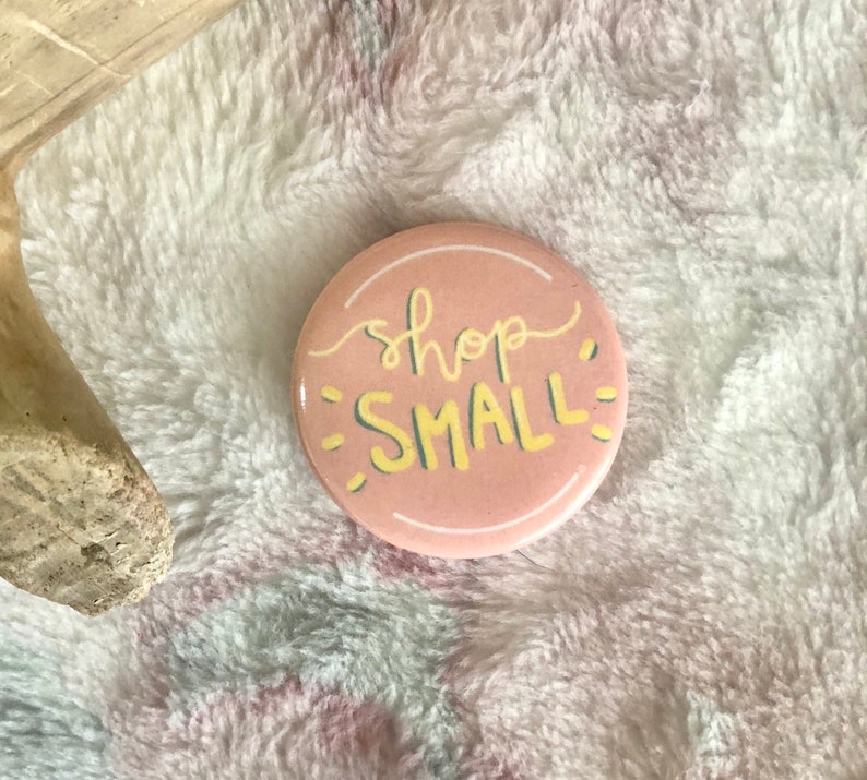 K&ouml;nnte beinhalten: Ein rosa Knopf mit dem Text "shop small" in gelber Schrift mit gr&uuml;nem Umriss.