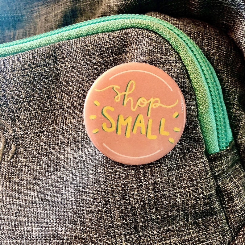 K&ouml;nnte beinhalten: Ein rosa Knopf mit dem Text "shop small" in gelber Schrift. Der Knopf befindet sich auf einem grauen Stoffhintergrund.