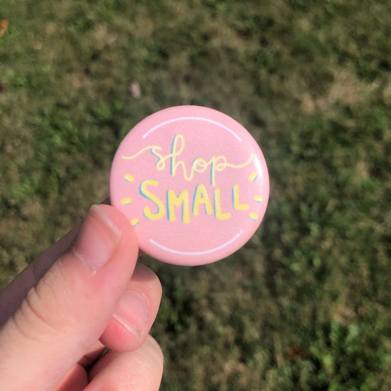 K&ouml;nnte beinhalten: Rosa Knopf mit dem Text "shop small" in gelber und blauer Schrift.