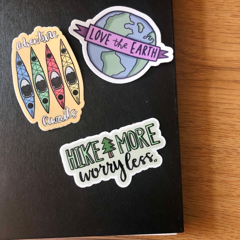 May include: Three colorful stickers with outdoor adventure themes. The first sticker features four kayaks in a row with the text "adventure awaits". The second sticker features a green, blue, and white globe with a pink banner that says "LOVE the EARTH". The third sticker features a green tree with the text "HIKE MORE worry less."