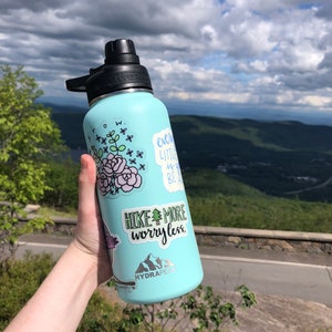 May include: A light blue reusable water bottle with a black lid and several stickers. The stickers include a floral design, a quote that says "every little thing is gonna be alright", a quote that says "hike more worry less", and a logo for the brand "Hydrapeak".