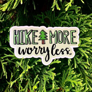 May include: A white sticker with green text that reads "Hike More Worry Less" with a small green tree illustration in the middle. The sticker is on a green leafy background.
