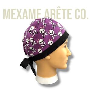 May include: Purple scrub hat with a skull and crossbones pattern and pink bows. The hat has a black elastic band and ties at the back.