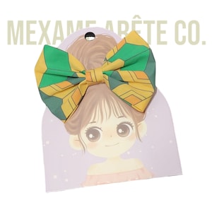 Puede incluir: Un lazo verde y amarillo con un patr&oacute;n geom&eacute;trico. El lazo est&aacute; en una tarjeta con una ilustraci&oacute;n de dibujos animados de una ni&ntilde;a con cabello casta&ntilde;o.